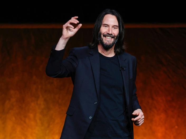 Keanu Reeves, wearing all black, smiles with his hand in the air while onstage at CinemaCon in April 2022.