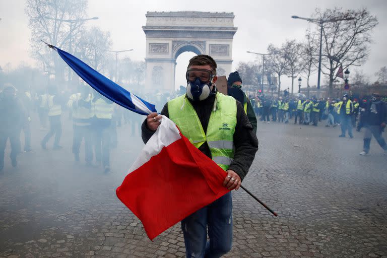 Un manifestante con un chaleco amarillo sostiene una bandera francesa mientras camina entre gases lacrimógenos en la avenida Champs-Elysees, cerca del Arco del Triunfo, durante un día nacional de protesta por el movimiento de "chalecos amarillos" en París, Francia, 8 de diciembre de 2018