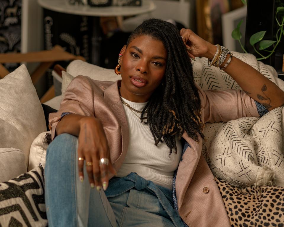 Interior designer Carmeon Hamilton poses on a couch, wearing a blush pink blazer over a white shirt with blue jeans. She's wearing rose gold and silver jewelry, including rings, bracelets, and a watch.