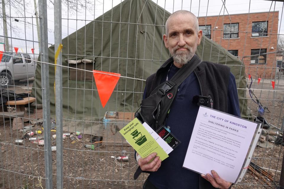 Kirk Sabiston stands in front of the tent he's currently living in at Kingston's Belle Park. He's holding information provided by the city on March 14, 2023, outlining its plans to begin forcing encampment residents to pack up their shelters each day starting next month.