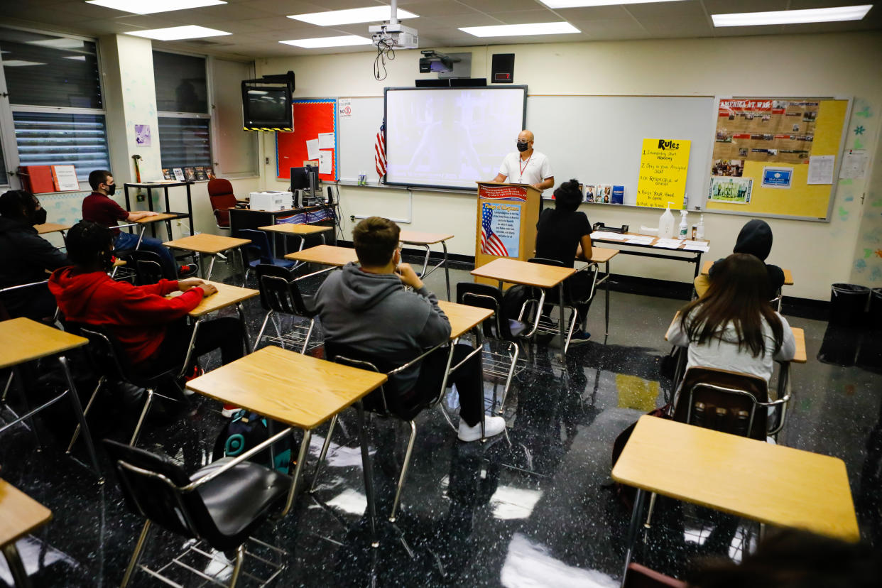 Students sit at their desks while a teacher stands at the front of the classroom at a podium.