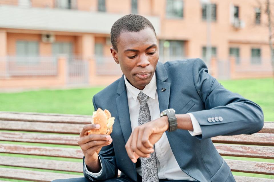 african american businessman eating a hamburger, watches wristwatch sitting on a bench in the park