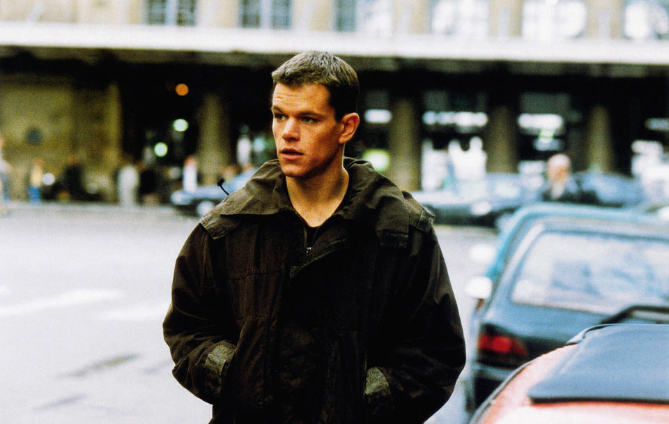 Matt Damon wearing a dark jacket stands on a city street with his hands in his pockets. Buildings and cars are in the background, in a scene