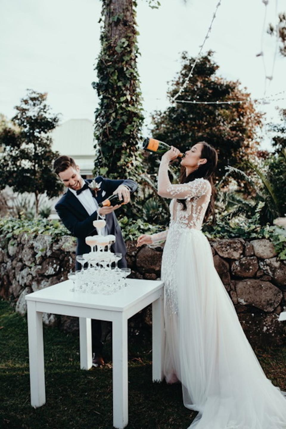 A bride drinks champagne as her groom pours another bottle of champagne into a champagne tower.