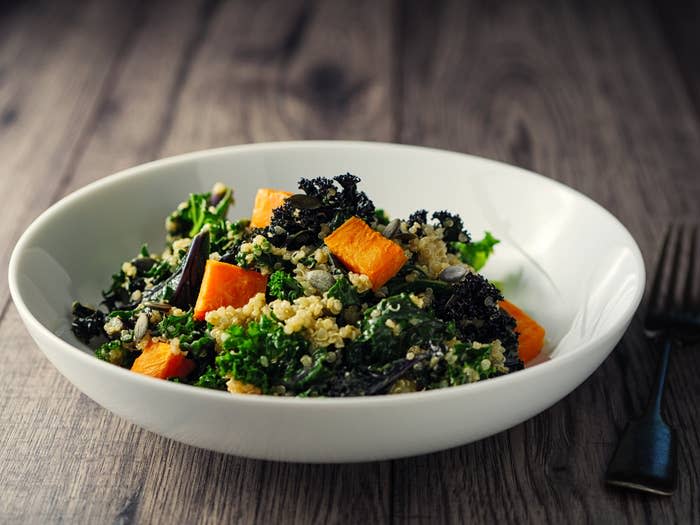 A bowl of quinoa salad with kale, roasted butternut squash, and various seeds placed on a wooden table next to a fork