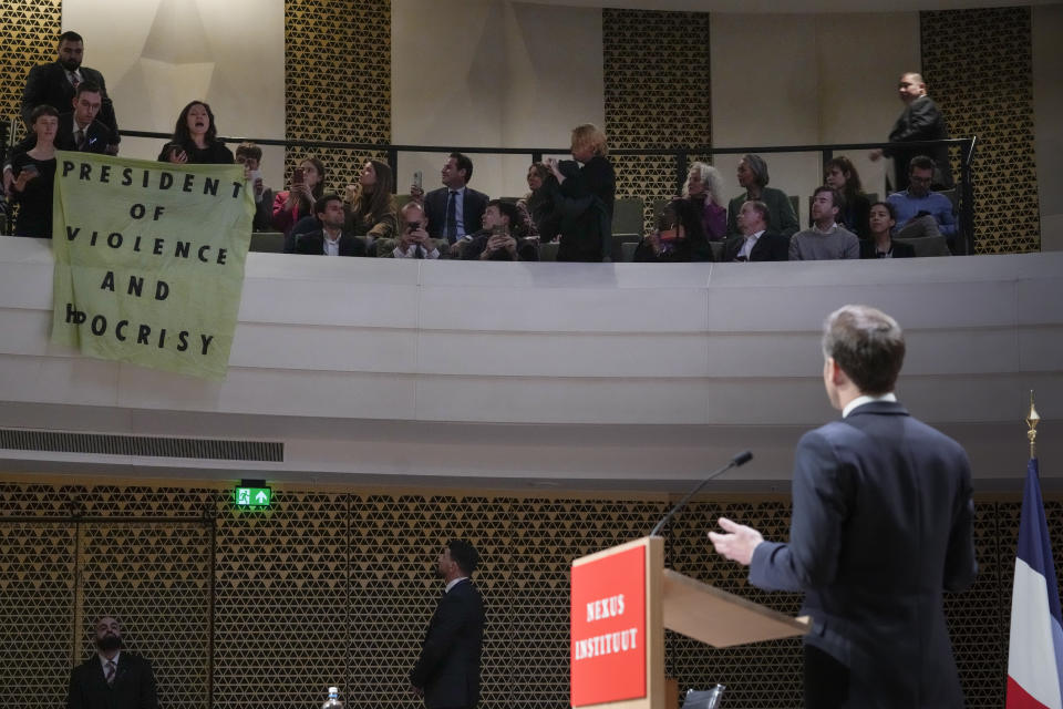 French President Emmanuel Macron, right, looks at demonstrators unfolding a banner reading "President of Violence and Hypocrisy" as he explains his vision on the future of Europe during a lecture in a theatre in The Hague, Netherlands, Tuesday, April 11, 2023. (AP Photo/Peter Dejong)