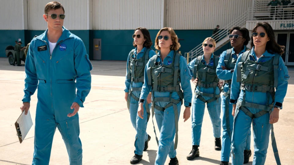 A man in a blue pilot jumpsuit leads a group of five women in similar suits