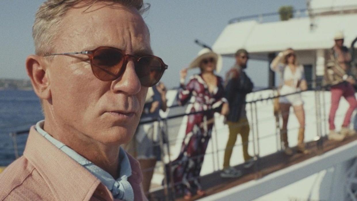 Daniel Craig in sunglasses, people behind him on a bridge to a boat