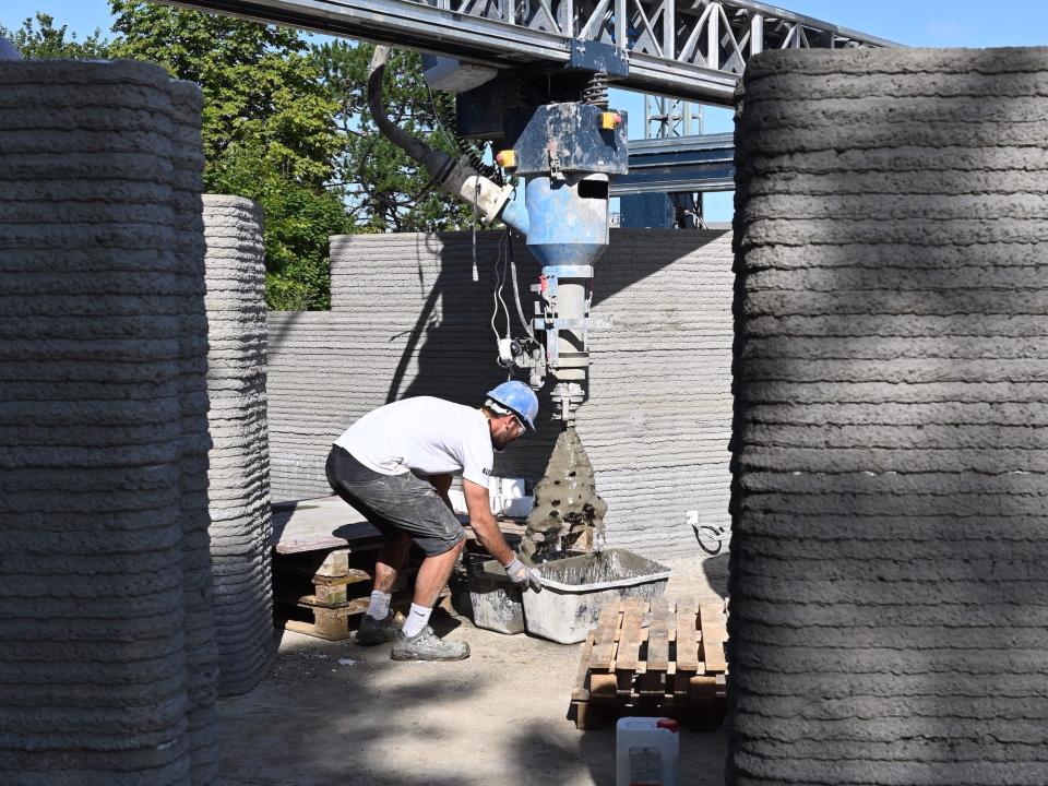 A 3D printer as it prints the walls of a tiny home. Someone is in the home doing construction.