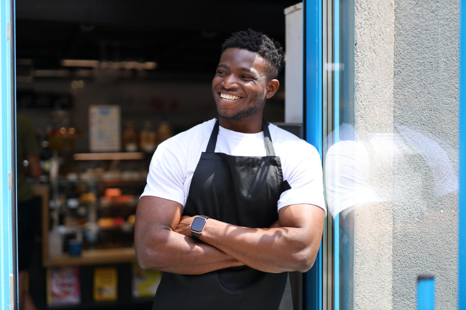 Successful black man who is wearing an apron and standing in front of a door. He is a young and confident owner, entrepreneur or worker who is smiling, looking or working for his small business.