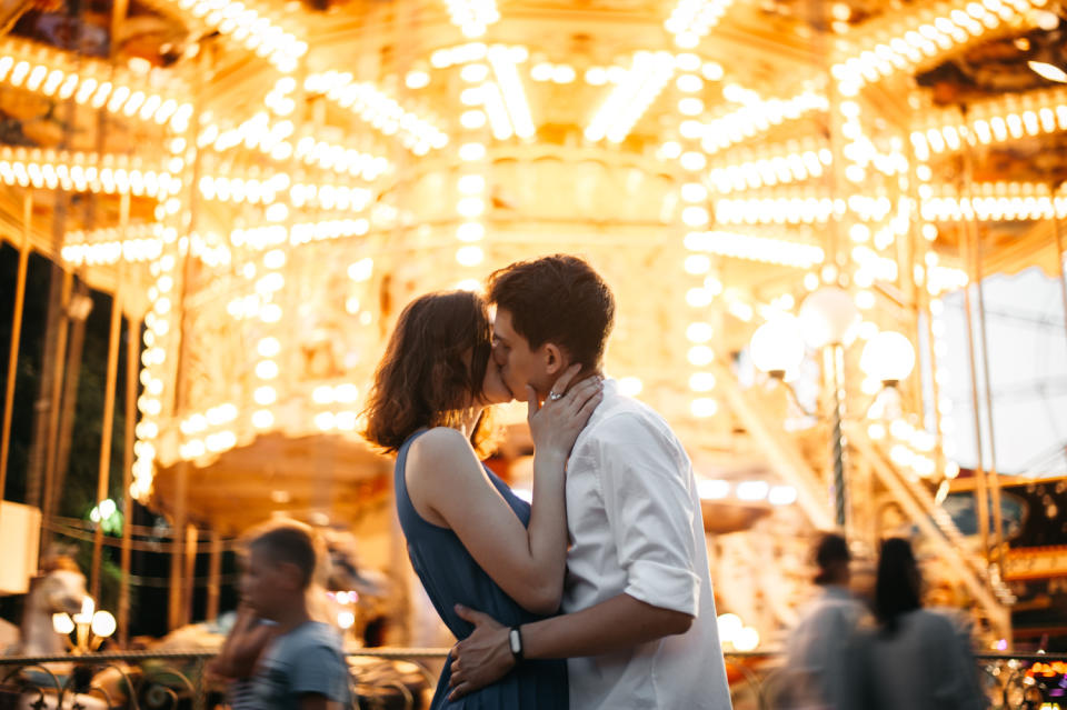 A couple kissing in front of a brightly lit merry-go-round. (Photo: Getty Images)
