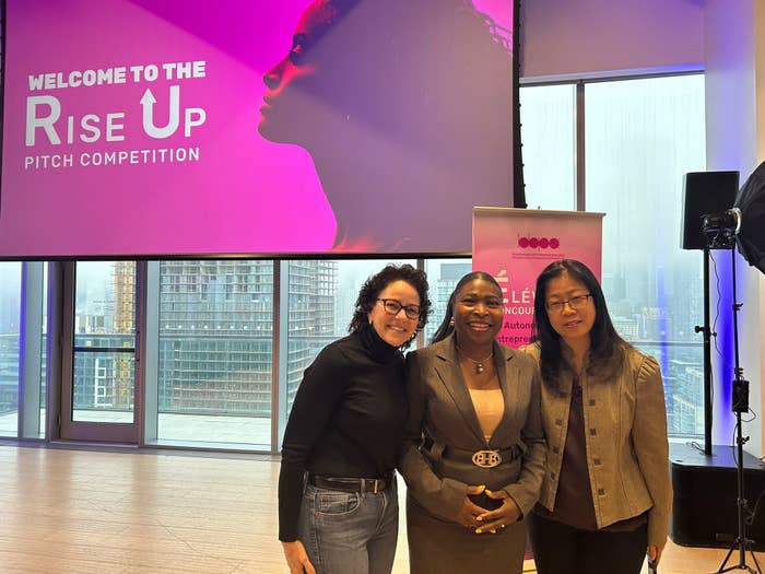 Three women smiling at the 'Rise Up Pitch Competition' event with cityscape background