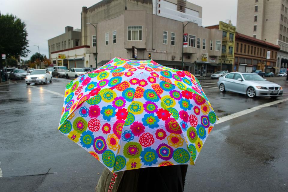 A woman waits for the light to change under a colorful umbrella on Market Street and San Joaquin Street in downtown Stockton. The bright colors of the umbrella against a drab background helps to draw the viewer's attention.