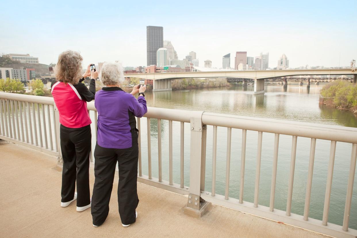 Two women standing on the 16th Street Bridge taking photos of downtown Pittsburgh on their cell phones.