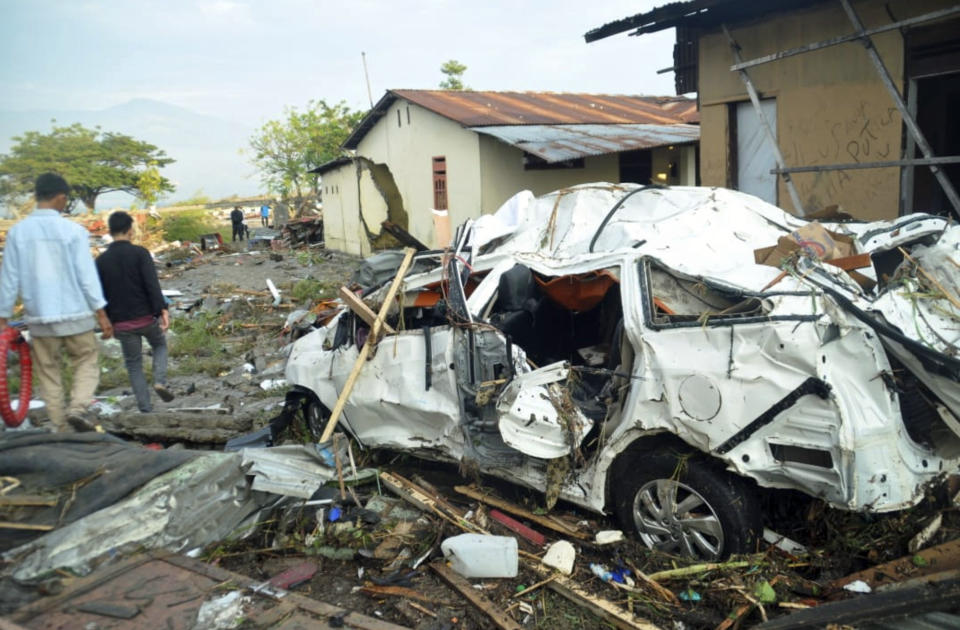 Indonesian men walk past the wreckage of a car following earthquakes and a tsunami in Palu, Central Sulawesi, Indonesia, Saturday, Sept. 29, 2018. A tsunami swept away buildings and killed large number of people on the Indonesian island of Sulawesi, dumping victims caught in its relentless path across a devastated landscape that rescuers were struggling to reach Saturday, hindered by damaged roads and broken communications. (AP Photo/Rifki)