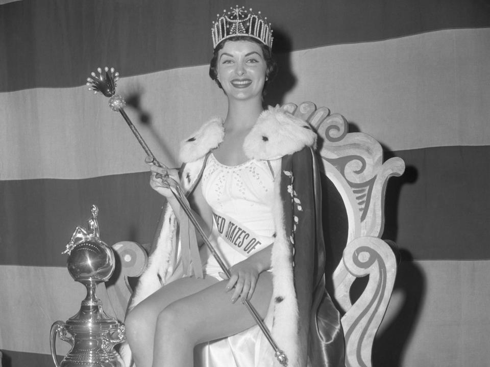 Miss USA 1957 Leona Gage sits on a throne with a tiara, scepter, and robe.