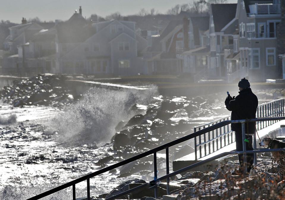 A man stops to take a photo of the surf pounding the shoreline in North Scituate at high tide on Tuesday, Jan. 4, 2022.