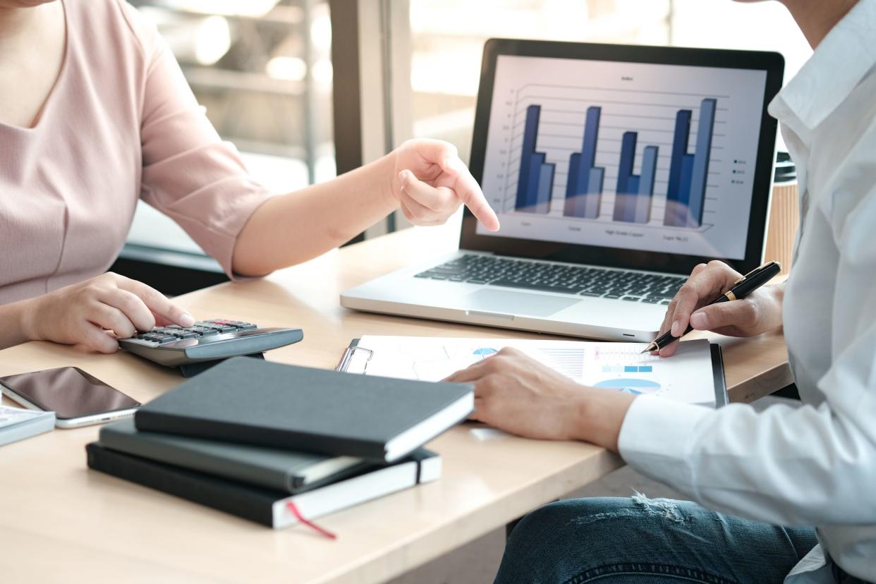 Focus on male and female financial managers' arms and hands at a desk with a laptop, female using calculator and male has a clipboard with blurred background of sunlight coming into windows