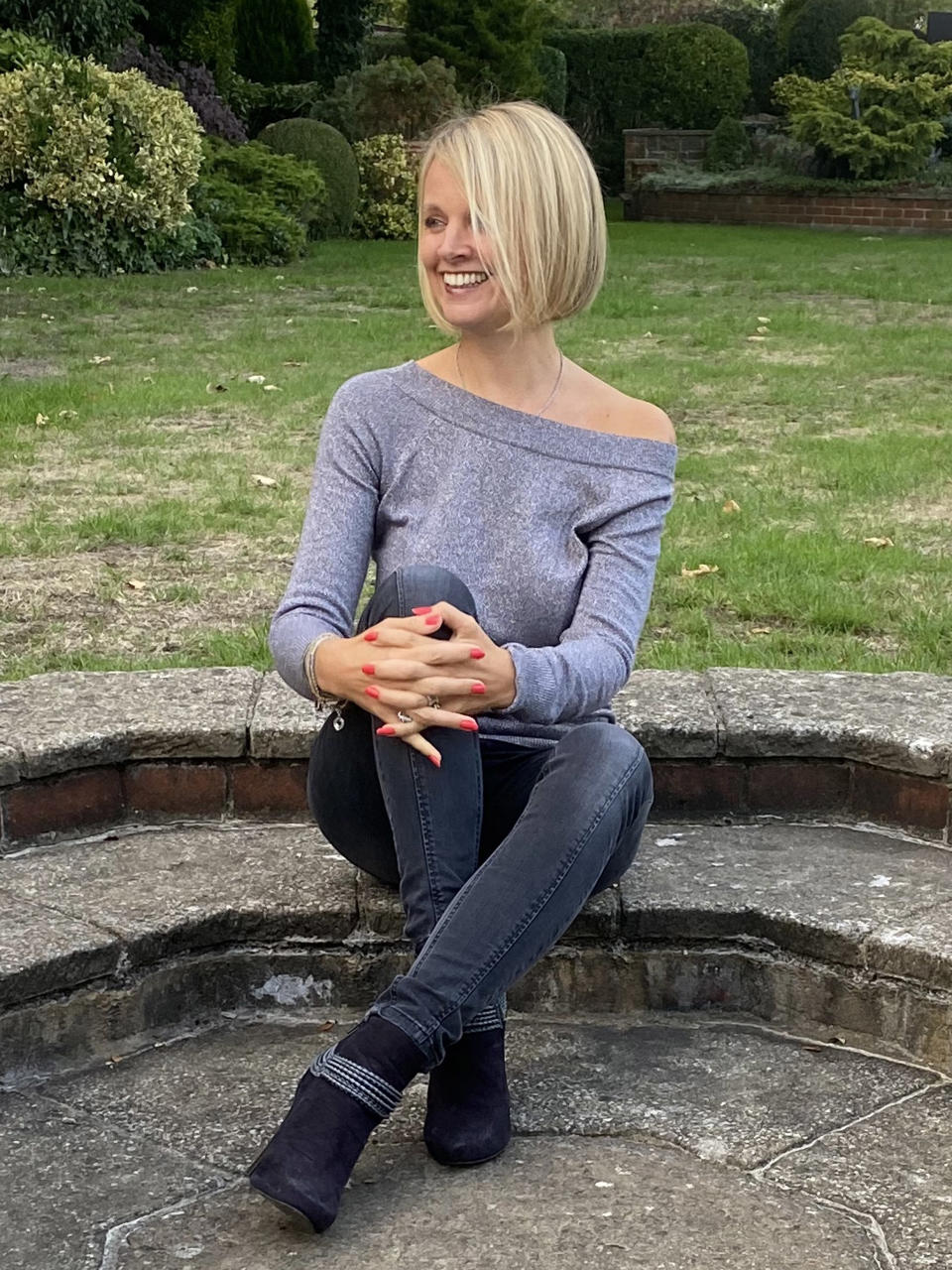 A blonde woman wearing a grey top and jeans sits on a stone step in a garden