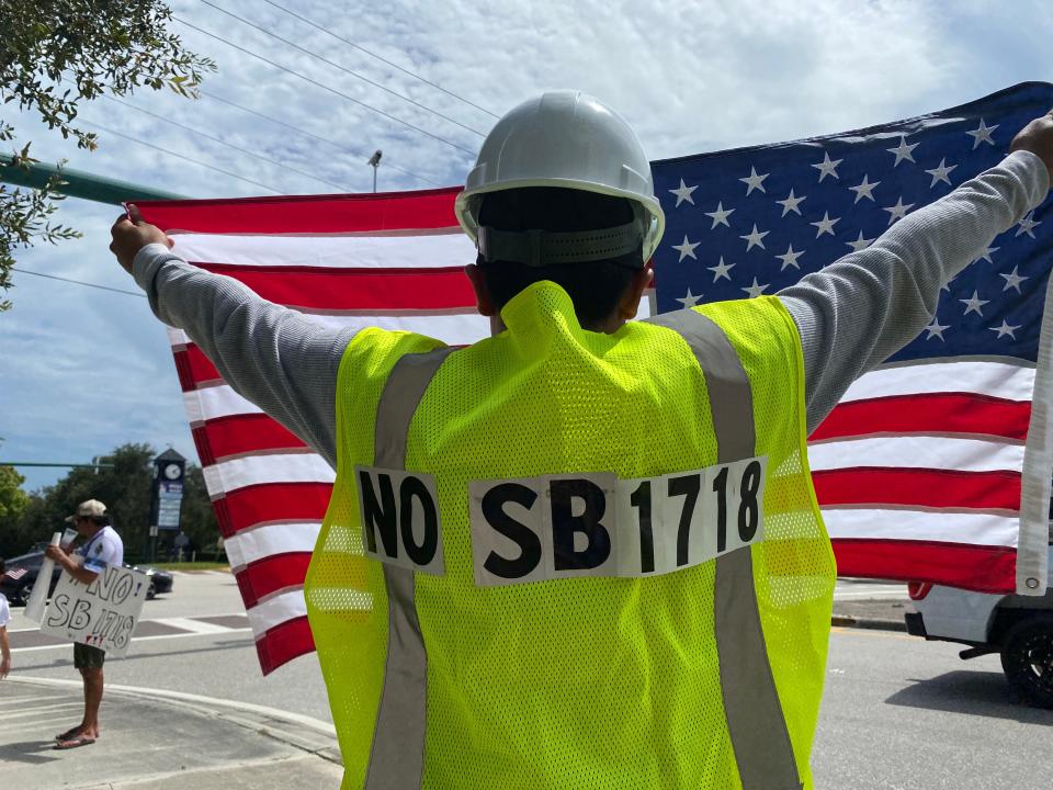 A protester at Indiantown Road and Military Trail in Jupiter holds up an American flag on Saturday June 24, 2023. His construction jacket bears a message against Senate Bill 1718, which imposes new restrictions on employers hiring immigrant workers.