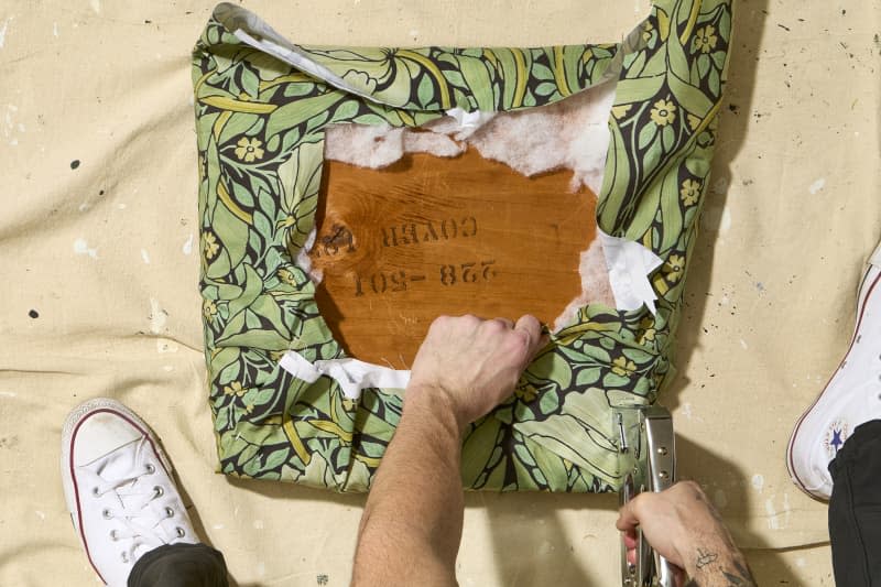 overhead shot of someone using a staple gun to staple the fabric to the bottom of the seat cushion.