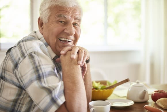 Smiling senior man seated at a kitchen table, hands folded under his chin, with food and dishes on the table in front of him.