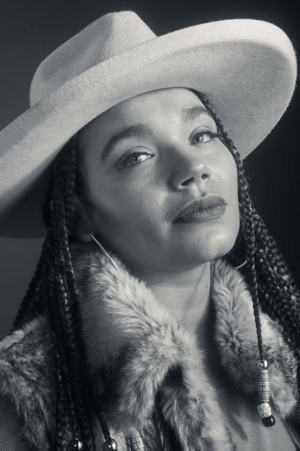 A woman in braids, a hat and a furry vest smiles.