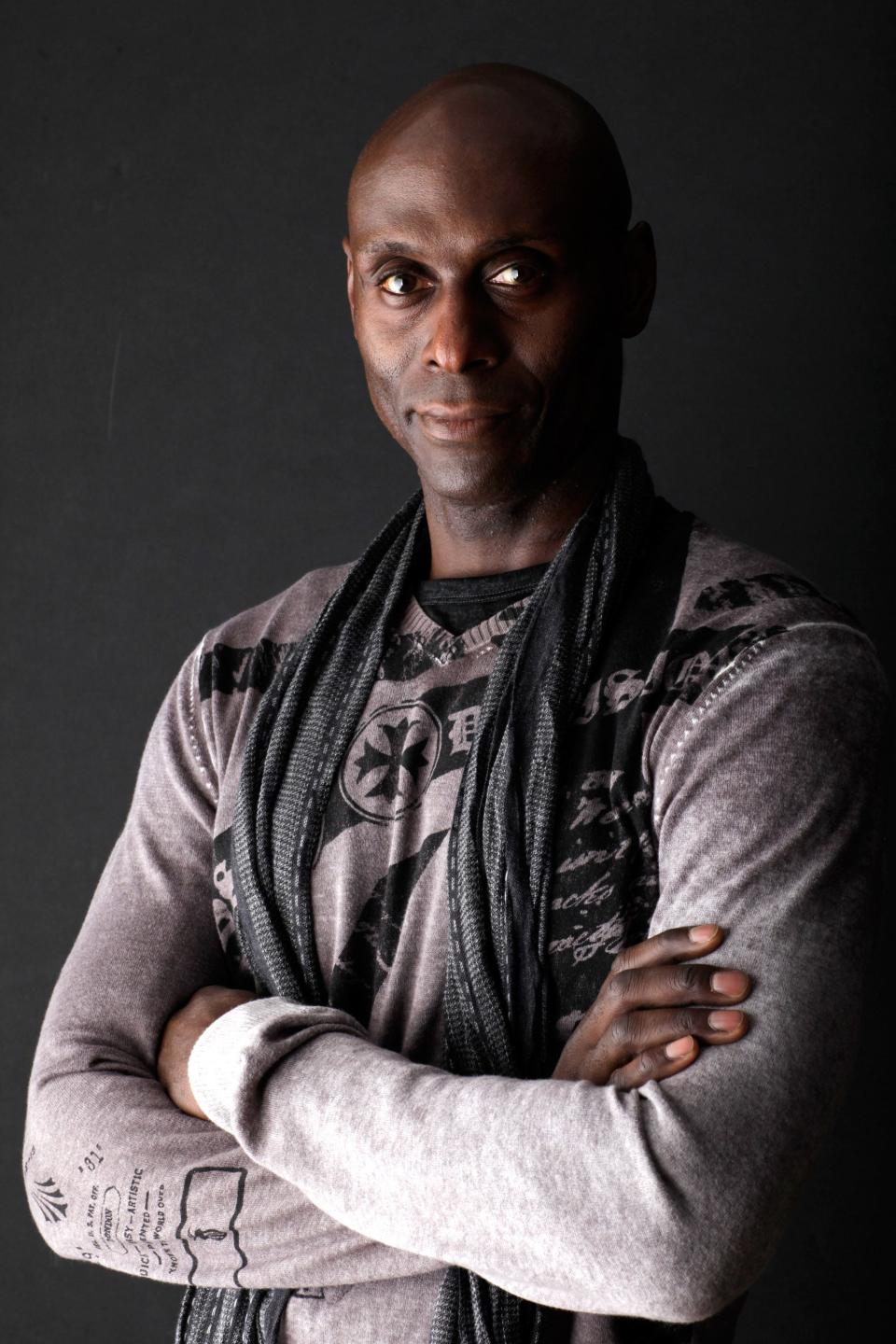 Lance Reddick in sweater and scarf with arms crossed