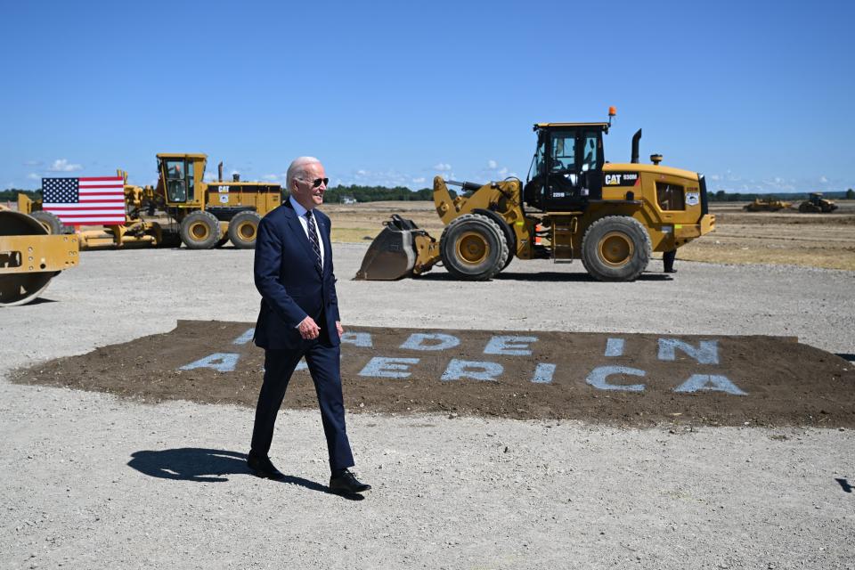 President Biden walks in front of a patch of dirt that says "Made in America." Behind him are construction vehicles and an American flag.