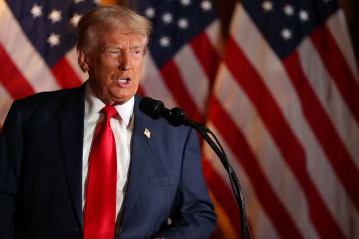 Donald Trump speaks at an event, standing in front of American flags and wearing a suit, shirt and tie