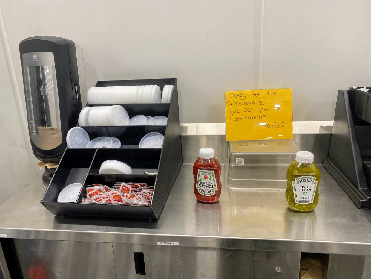 Condiments area for the cafe at Sam's Club, napkins, drinks top, salt, ketchup, and sweet relish, a sign explaining that Sam's Club is out of condiments, clean stainless steel table, white wall