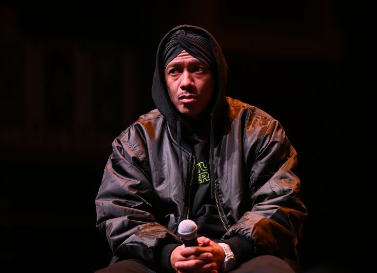 Nick Cannon sits onstage in a hoodie and leather jacket, holding a microphone on his lap with both hands.