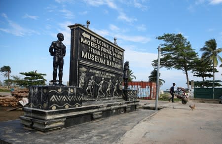 Man uses a wheel barrow as he walks past a bas-relief of shackled slaves embedded on a plague advertising the Mobee Royal Family Original Slave Relics Museum in Badagry