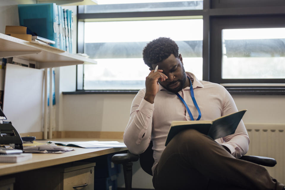 Man in an office sitting at a desk, focused on reading a book. Shelves with books and files are in the background