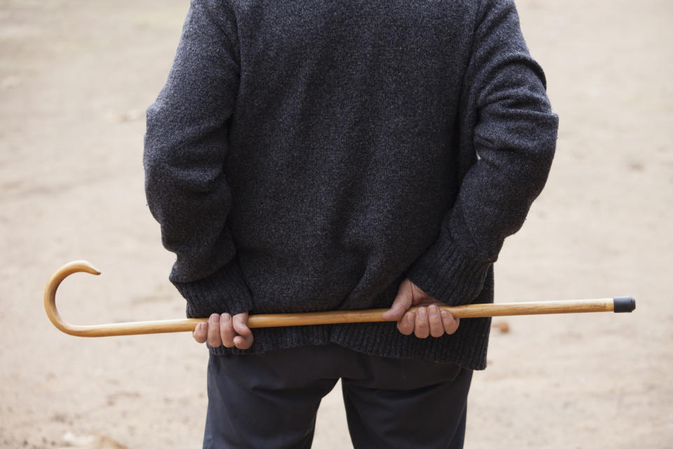 A person with their back turned holds a wooden cane behind them. The person is wearing a dark long-sleeve shirt and pants