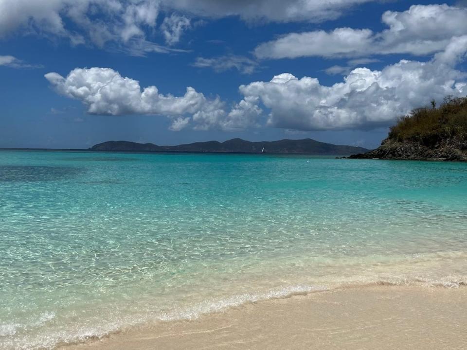 Low clouds over a beach with crystal-clear blue water. There are mountains in the background.