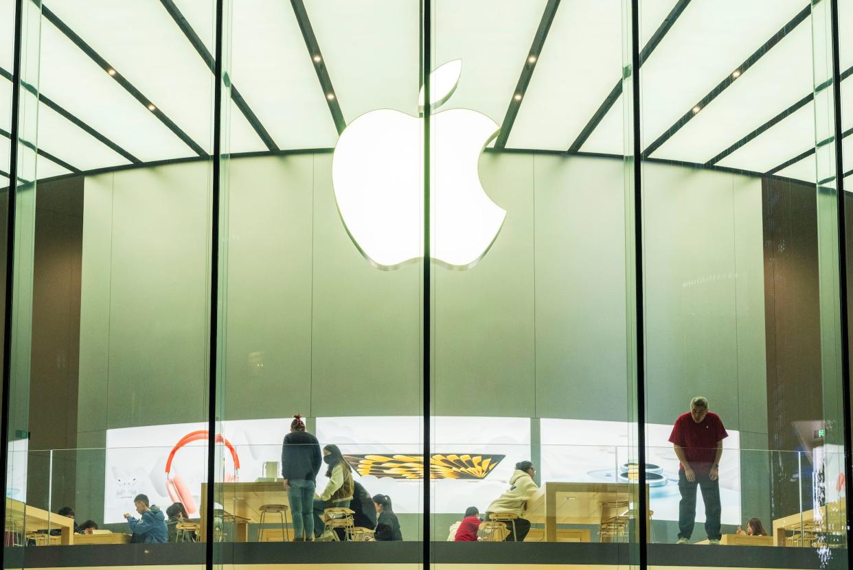 A large white Apple logo is illuminated above people sitting at wooden tables inside an Apple store