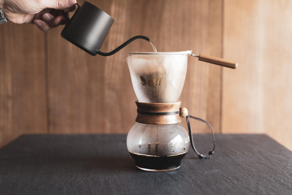 A hand pours hot water from a black kettle into a Chemex coffee maker with a wooden collar and leather tie, making fresh coffee