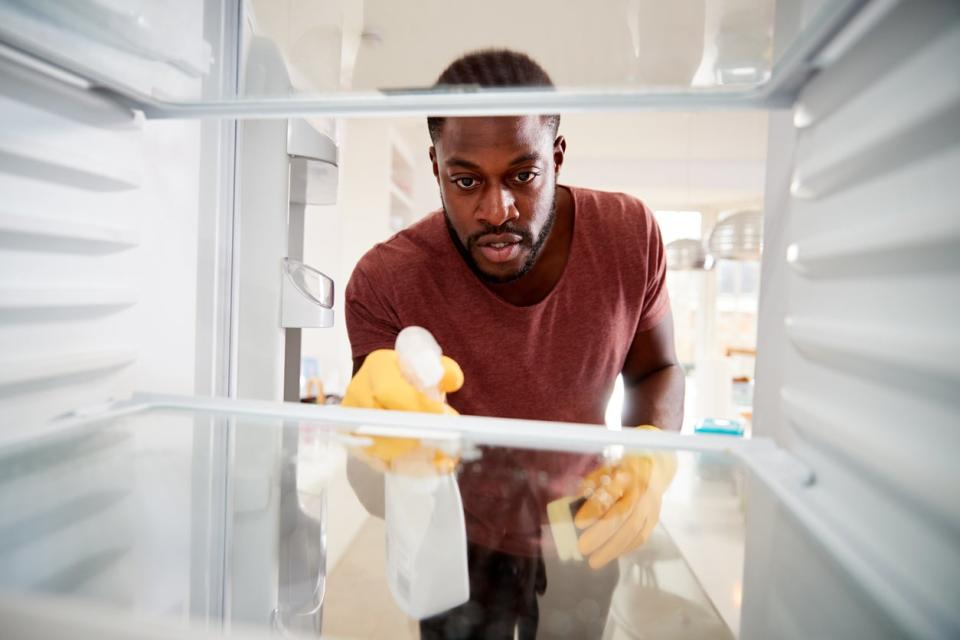 View looking out from inside an empty refrigerator as a man wearing rubber gloves cleans shelves.