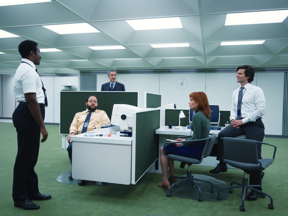 tramell tillman, zach cherry, john tuturro, britt lower, and adam scott in severance, sitting around a cluster of four desks in a brightly lit room with a green carpet