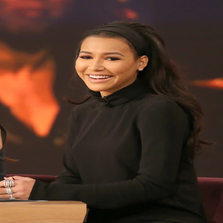 Naya Rivera, wearing a black dress, sitting at a desk on the set of The View