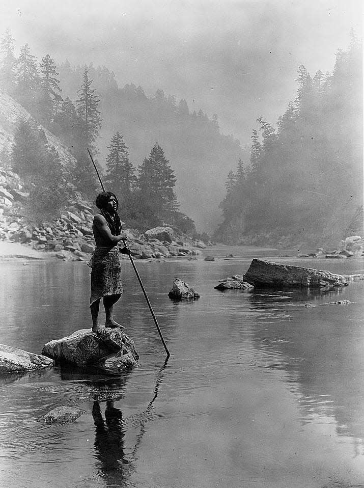 Hupa man with spear, standing on rock midstream, in background, fog partially obscures trees on mountainsides.
