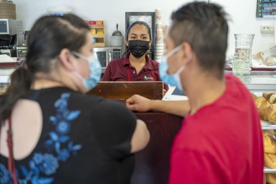 Two customers wearing masks wait at a cash register where Mayte Ramirez, a clerk, is also masked.