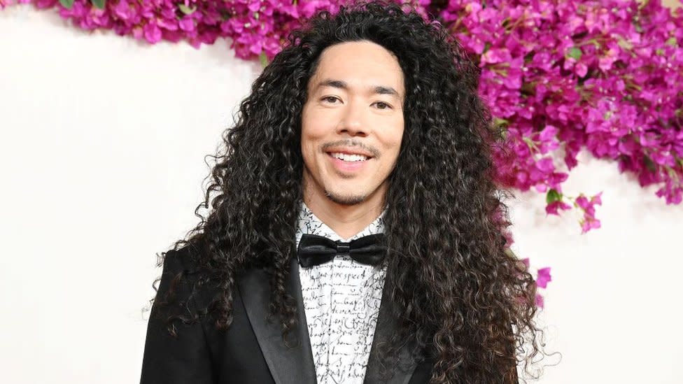 Cole Walliser at the 96th Annual Oscars held at Ovation Hollywood on March 10, 2024 in Los Angeles. Cole is a Canadian man in his 40s, with long curly brown hair past his shoulders. He has brown eyes and a short moustache. He wears a black tuxedo with a black bow tie and a white shirt with black script on it. He's pictured on the red carpet against a white backdrop with hundreds of pink flowers.