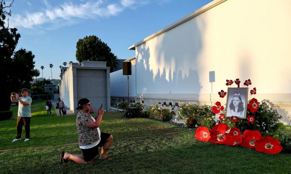 A man kneels in the grass to take a picture of Judy Garland's grave which is strewn with oversized red poppies. Other people can be seen in the background taking pictures.