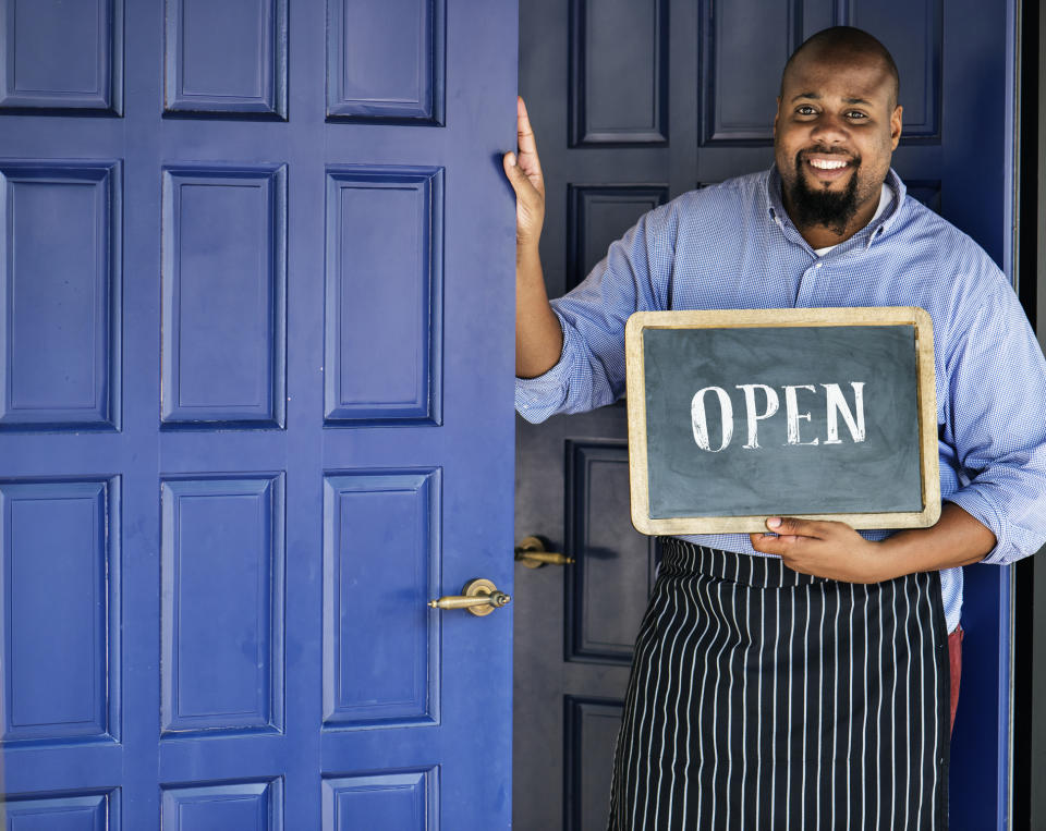 A happy cafe owner stands in an apron at the entrance to his business and holds a chalkboard sign which says "Open."