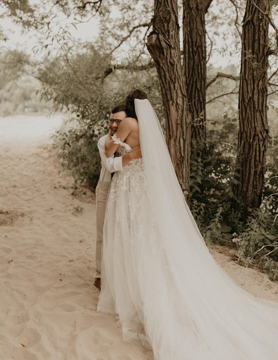 A bride and her brother hug on a beach in wedding attire.