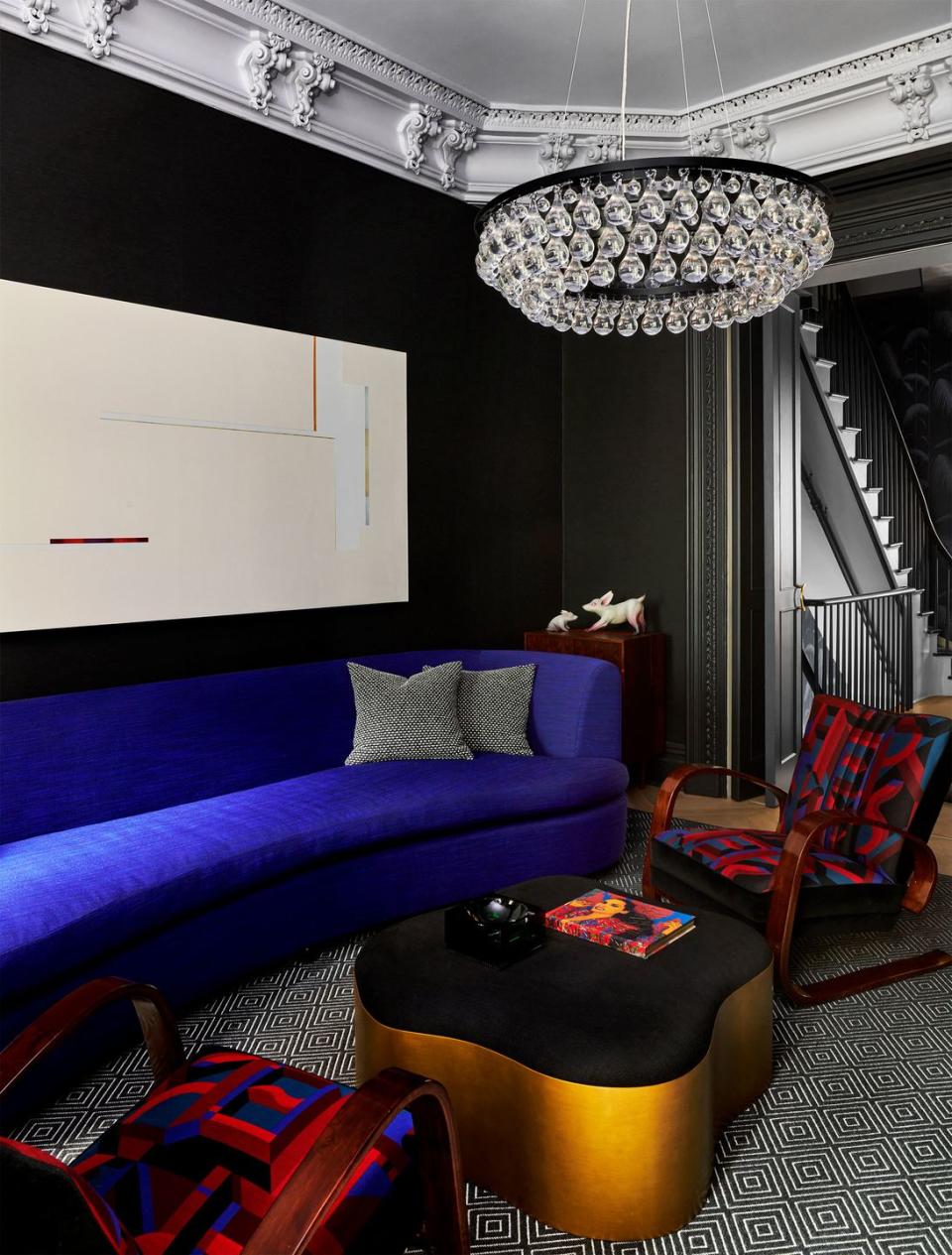 a living room has black walls with white ornate ceiling molding, a chandelier made of crystal teardrops, a cobalt blue sofa, a white artwork on the wall, a curved edge cocktail table, two patterned armchairs