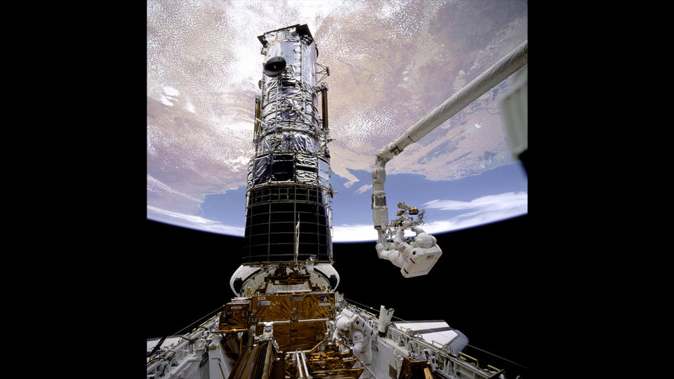 a large chrome cylinder (Hubble) is attached to the cargo bay of a space shuttle.  The Earth hangs above it, a mechanical area extends to the right.