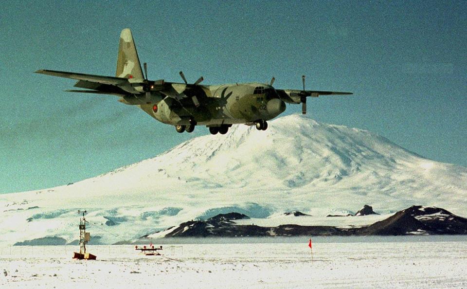 A large, camouflaged C-130 flying in the Antarctic with a snowy mountain in the background.